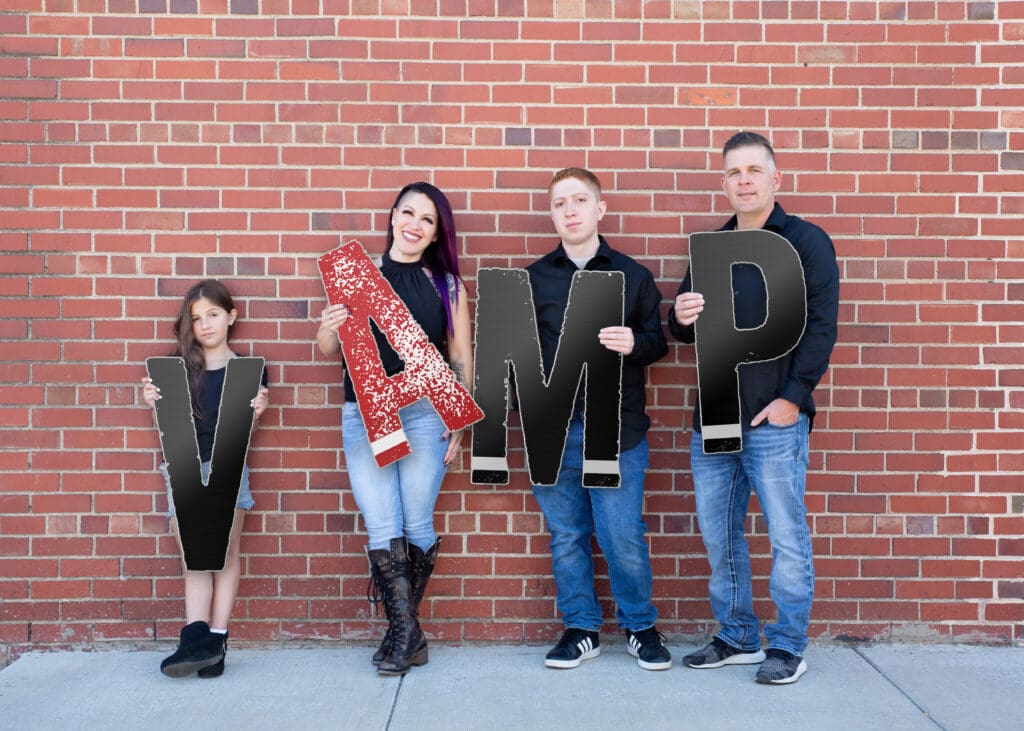 The VAMP Stucco family holding large letters spelling “VAMP” in front of a red brick wall.